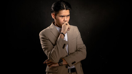 Asian man in beige blazer and white shirt posing with hand on mouth in thoughtful expression, standing against dark background, ideal for concepts like thinking, strategy, or decision-making