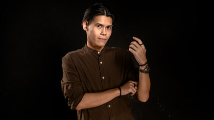 Asian man in brown button-up shirt posing in studio with dark background, looking at his raised...