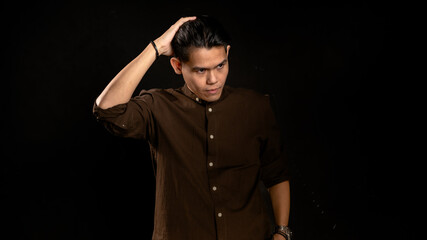 Asian man in dark brown shirt styling his hair with one hand while looking sideways in studio against dark background, perfect for fashion, grooming, confidence, or personal branding visuals