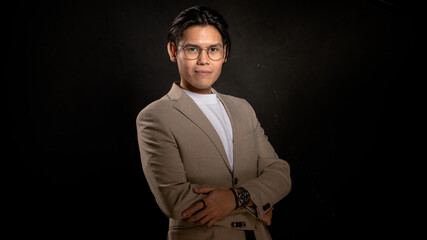 Asian man wearing beige blazer and white t-shirt poses in studio against dark background with hands...