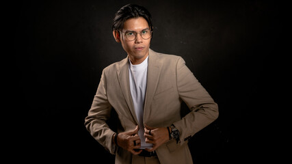 Asian man in beige blazer and white t-shirt adjusting jacket while looking sideways in studio with dark background, expressing confidence and style, ideal for business, fashion, or branding use