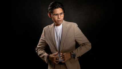 Asian man in beige blazer and white t-shirt adjusting jacket while looking sideways in studio with dark background, expressing confidence and style, ideal for business, fashion, or branding use