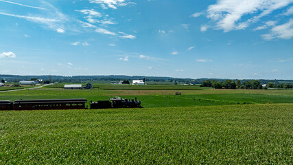 A vintage steam train travels through expansive green fields under a bright blue sky, surrounded by distant hills and farm buildings, creating a picturesque rural atmosphere.