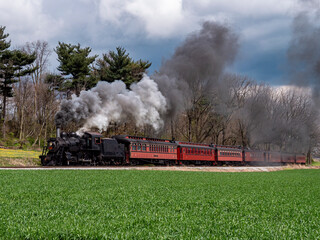 A vintage steam locomotive pulls a red passenger car along a scenic route. The train emits plumes of smoke against a backdrop of green fields and overcast skies, capturing a moment of nostalgia.