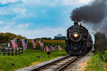 Obraz premium A classic steam train makes its way through a picturesque rural landscape, flanked by American flags. The scene captures the charm of vintage rail travel on a clear day.