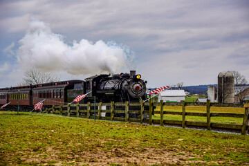 Obraz premium Historic steam locomotive pulls vintage passenger cars along tracks while puffs of steam rise against a cloudy sky. The setting features a green field and wooden fence nearby.