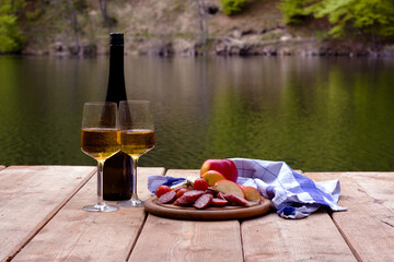 A scenic picnic on a wooden dock features wine, glasses, salami, apples, and a checkered cloth by a calm lake