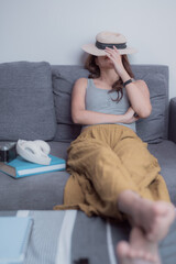 Obraz premium Young woman lying on a cozy sofa, hiding her face with a stylish straw hat, surrounded by books, a camera, and headphones, savoring moments of relaxation and tranquility at home