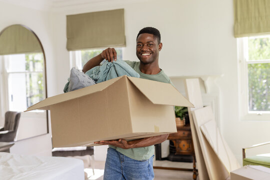 Smiling African American man carrying moving box inside bedroom at home, with blankets and pillows