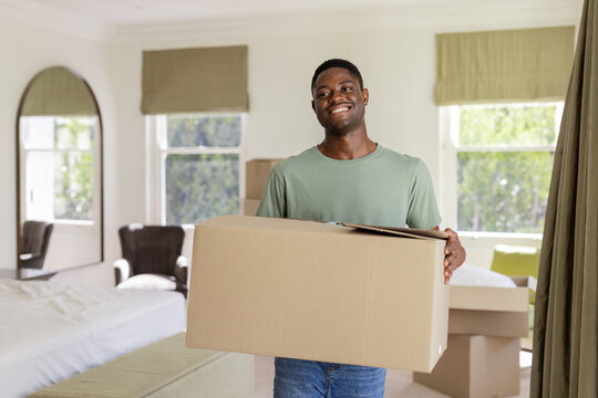Moving in, carrying african american man smiling and unpacking boxes in bedroom, including mirror - Powered by Adobe