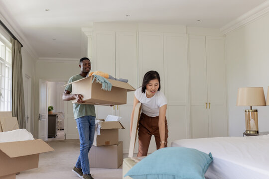 Diverse couple unpacking cardboard boxes in bright bedroom, with framed artwork and blankets