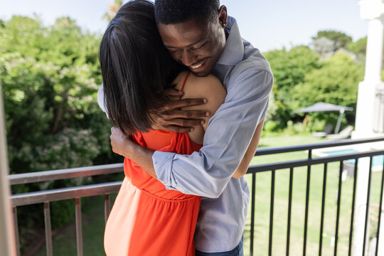 Hugging diverse couple standing on balcony overlooking backyard, with patio umbrella - Powered by Adobe