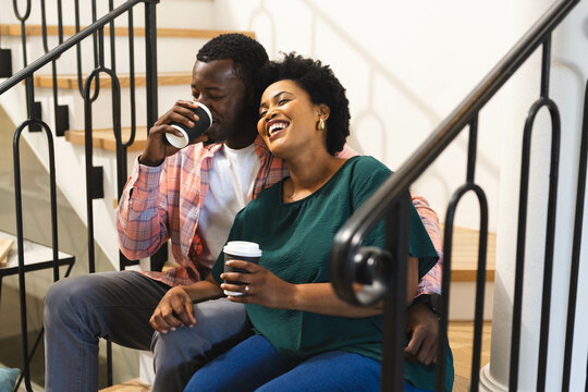 Laughing African American couple sipping coffee on staircase at home, with black metal railings