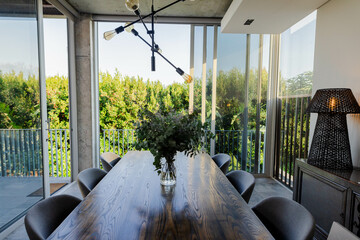 Long dining table is featuring glass vase with green foliage beside sliding doors in dining room