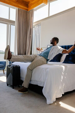 Relaxing African American man stretching legs on bed in hotel, with suitcase, windows, copy space