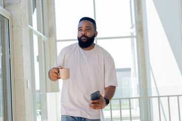 Standing mid-adult African American man checking smartphone in modern living space, with coffee mug