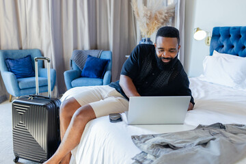 African American man typing on laptop in modern hotel room on bed, with smartphone and suitcase