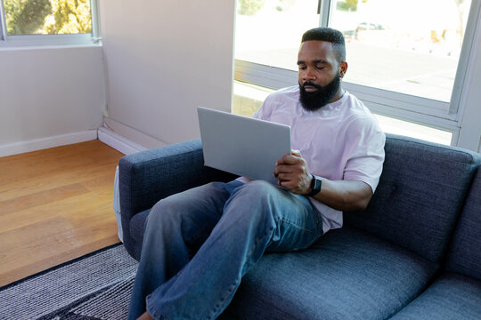 Working African American man typing on laptop on gray sofa in living room, with smartwatch
