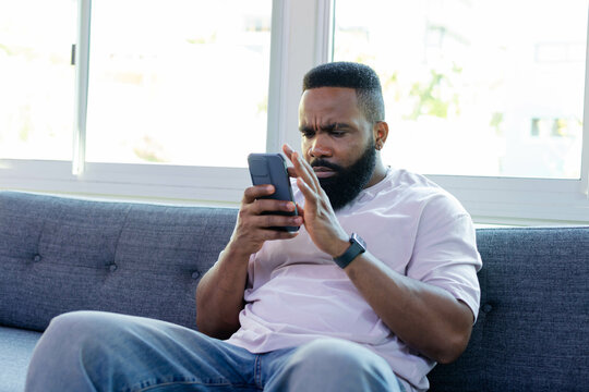 Sitting African American man viewing smartphone on gray couch by window, with smartwatch