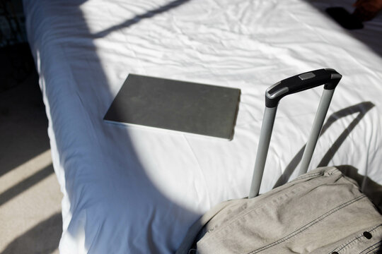 Closed silver laptop is resting on sunlit sheets while light gray suitcase is standing near window