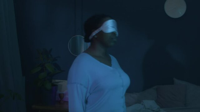 Slow motion shot of young African American woman in satin eye mask and pajamas standing in dark bedroom, moving from side to side while sleepwalking at night