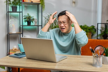 Displeased sad Asian man use laptop notebook typing browsing working, loses becoming surprised...