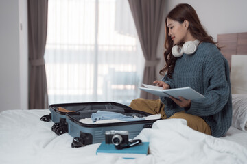 Young woman with headphones around her neck checking her travel journal while preparing luggage on bed in hotel room, getting ready for her next adventure