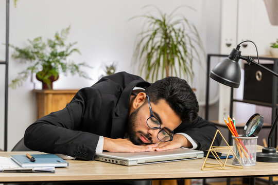 Bored sleepy Indian businessman worker working on laptop computer, leaning on hand falling asleep at office table. Exhausted tired freelancer workaholic young man. Employment, occupation, workless
