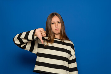 Young brunette Hispanic teenager wearing a striped sweater looking unhappy and angry, showing rejection and negativity with a thumbs-down gesture, isolated over blue background. Bad expression.