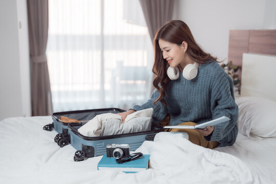Young asian woman packing clothes in suitcase on bed, checking her travel itinerary and preparing for vacation, she is smiling and getting ready for her trip