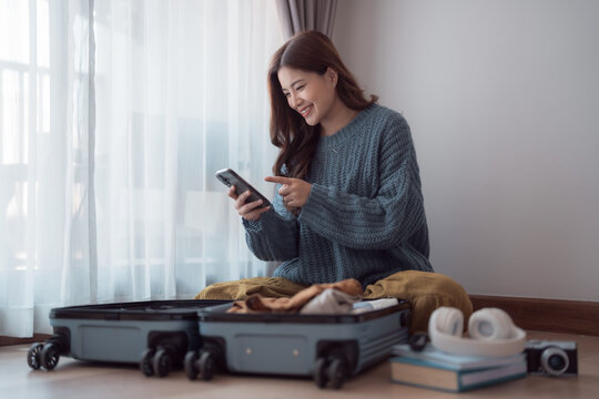 Happy young woman packing clothes in suitcase and using smartphone, checking her travel itinerary or online booking information while preparing for vacation trip