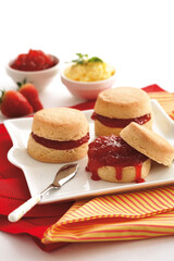 Three scones with jam are on a white plate. There are bowls of jam and cream, and strawberries nearby.