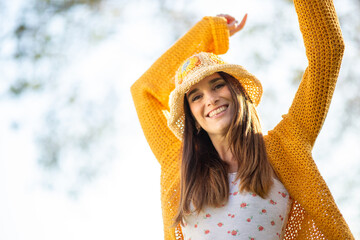 Joyful woman in a yellow cardigan and a hat raising her arms under the sunlight
