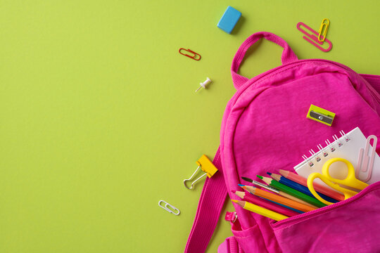 Creative arrangement of a pink backpack filled with school essentials, including pencils, scissors, a notebook, and more, set against a green background to evoke educational concept