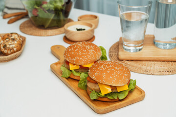 Focusing on double giant fried meat hamburger meal on square handle wooden board. Decorated by salad, homemade bun bread, beside drinking water on fast healthy food concept on white table. Infobahn.