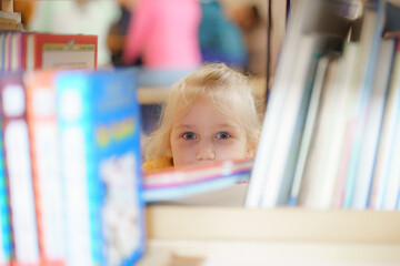 Child exploring a library shelf while curious peers engage in activities around her during a school afternoon