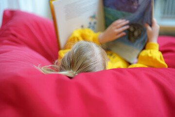 Child reading book while relaxing on soft red bean bag in comfortable indoor space during cozy afternoon