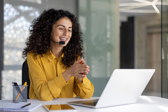 A cheerful customer service representative wearing a headset smiles while working on her laptop. The woman is in a modern office.