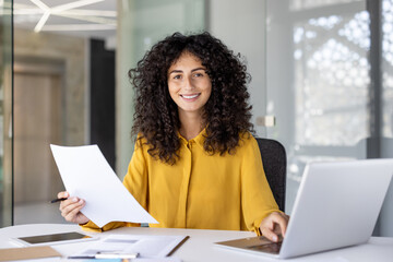 A smiling woman in a yellow blouse sits at her desk, holding a document. She is looking directly at the camera. A laptop sits in front of her.