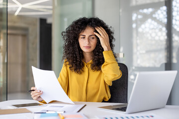 A concerned woman reviews documents in her office, appearing stressed while working on her laptop and analyzing papers at her desk.