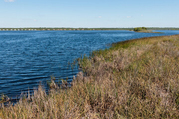 The marshy shoreline of Lake Shelby in the foreground, the cabins along the distant shore within Gulf State Park, Gulf Shores, Alabama in mid-April