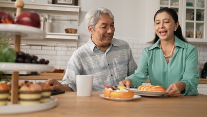 Smiling grandfather with casual cloth sitting at table while grandmother presenting breakfast. Senior couple talking and spending time or supporting together. Healthy relationship concept. Myrmidon.