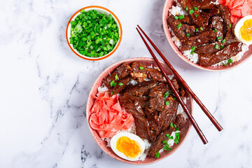 Gyudon Japanese beef with rice, pickled ginger, boiled egg slice, garnished with green onions, in a bowl with chopsticks,