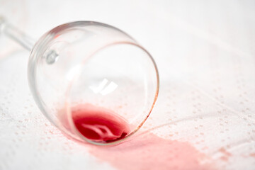 Empty wine glass lies on its side, spilling red liquid onto crisp white tablecloth. Scene captures moment of accidental spill, highlighting stark color contrast and casual atmosphere