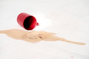 Red mug lies on white fabric, spilling coffee and creating dark stain. Bright lighting accentuates contrasting colors and highlights mess. Image evokes sense of accident and urgency for cleaning