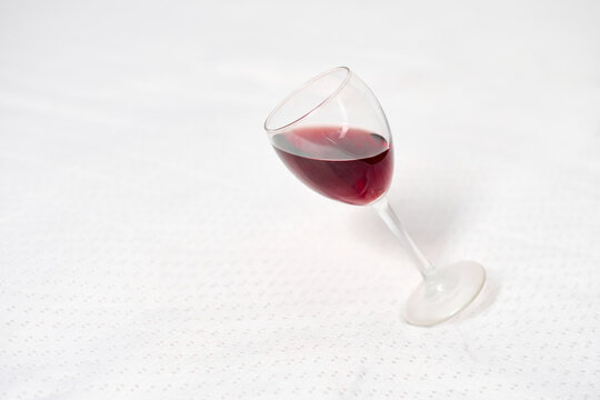 Wine glass with red liquid rests tipped on white tablecloth. Subtle lighting enhances contrast between dark wine and bright background, creating minimalist, elegant scene