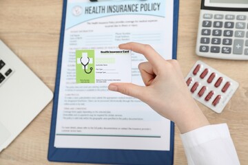 Obraz premium Doctor holding health insurance card above policy form at wooden table, closeup