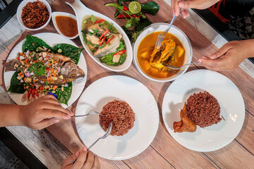 Overhead view of hands sharing a traditional Thai food meal with fried fish, spicy soup, yellow curry, and brown rice, Authentic Cuisine.