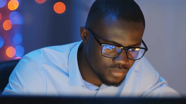 Office worker with glasses focusing on computer screen, illuminated by its light, with blurred colorful lights in the background, suggesting late night work