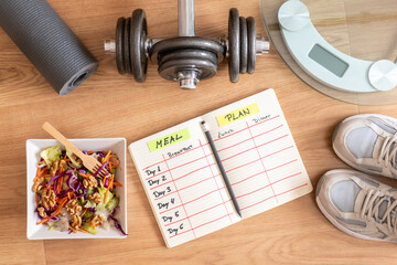 Bird's-eye view of a wooden floor featuring a healthy salad, meal planning notebook, dumbbell,...